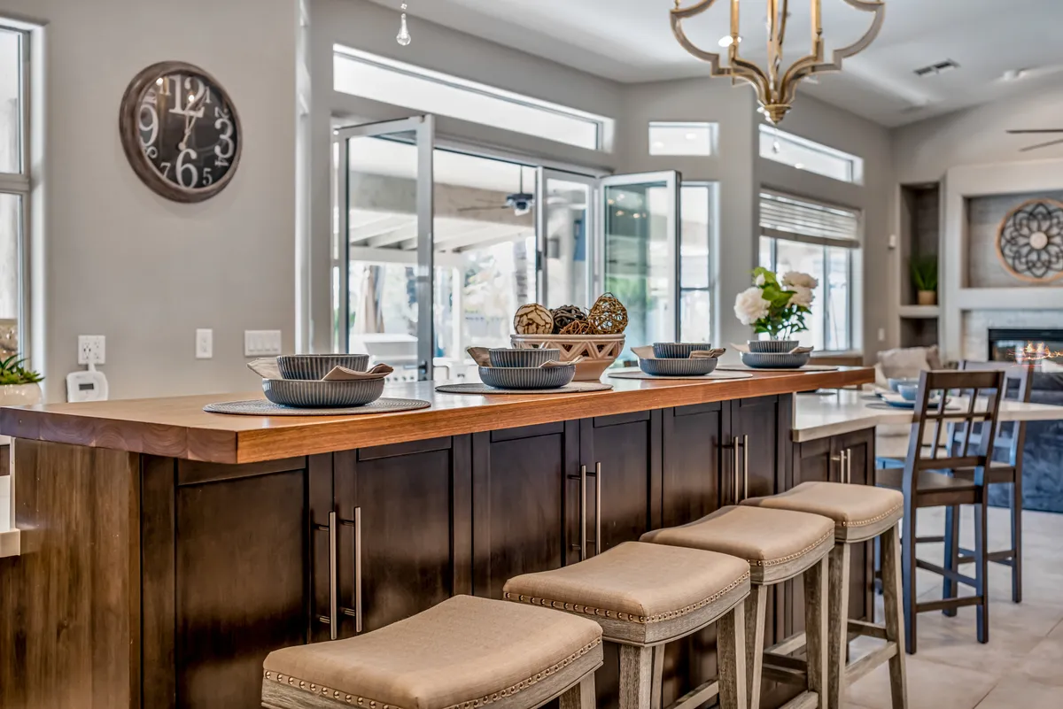 A stylish kitchen island stands at the center, surrounded by bar stools and topped with decorative bowls. A modern chandelier hangs overhead, while a clock adorns the wall. The space opens to the adjacent living and dining areas, highlighting the open-concept flow and inviting spaces for casual dining or entertaining.
