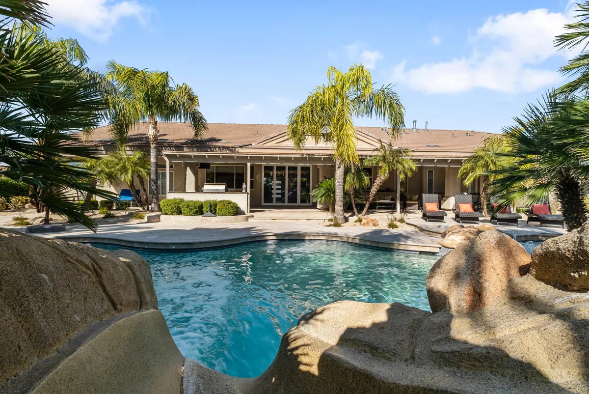 A wide view of the backyard reveals a sparkling swimming pool with an exciting sliding rock feature, surrounded by palm trees and a spacious patio. The area is perfect for both sunbathing and outdoor dining, emphasizing opportunities for family gatherings and fun.