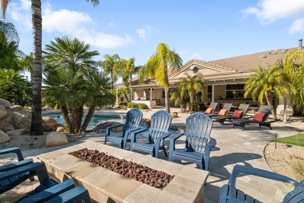 A vibrant exterior shot featuring a swimming pool flanked by deck chairs, a fire pit, and tall palm trees. The scene showcases the villa at dusk, with the fire pit ready for evening gatherings and the pool inviting for a refreshing dip or sunset swim.