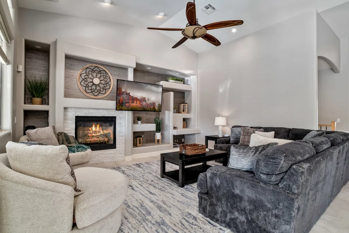 A cozy living room features a fireplace as its focal point, with plush sofas arranged for conversation and a TV unit above the mantel. Soft lighting and a ceiling fan create a comfortable atmosphere ideal for relaxing or storytelling.