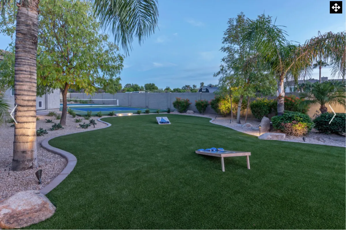 A spacious backyard is shown with a lush green lawn, a sparkling swimming pool edged by palm trees, a landscaped garden, and a visible cornhole game set on the grass. There's ample space for outdoor activities and family gatherings, and the warm sunset lighting enhances the inviting, playful mood.