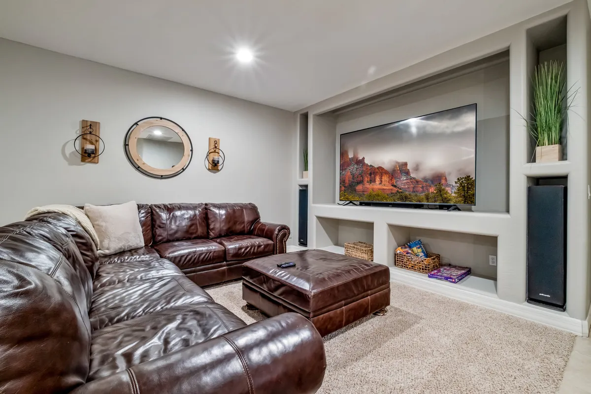 A comfortable interior living area featuring a large sectional sofa, a big flat-screen TV, a decorative mirror, and a textured ottoman. The mood is relaxed and perfect for movie nights or unwinding after a day of games.