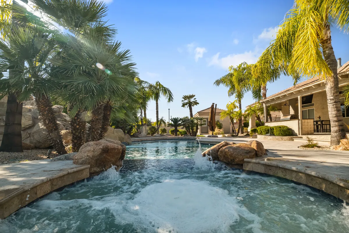 A luxurious outdoor pool framed by palm trees with a gentle water feature, surrounded by comfortable patio seating and lush landscaping. The setting appears ideal for both daytime lounging and evening social events, offering versatility for different types of gatherings.
