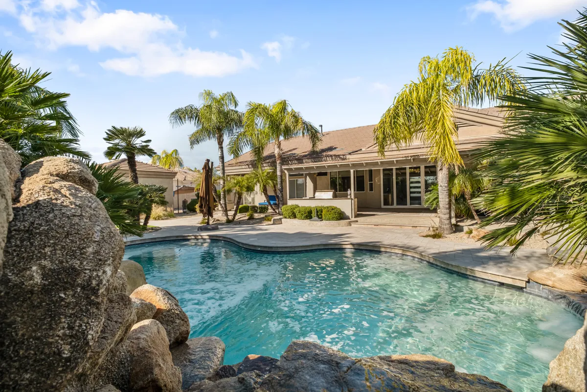 A tranquil backyard view featuring a large swimming pool framed by lush palm trees, natural rock formations, a generous patio area, and the beautiful facade of the house. The scene suggests ample space for swimming, sunbathing, and group activities, with a family-friendly, welcoming vibe.