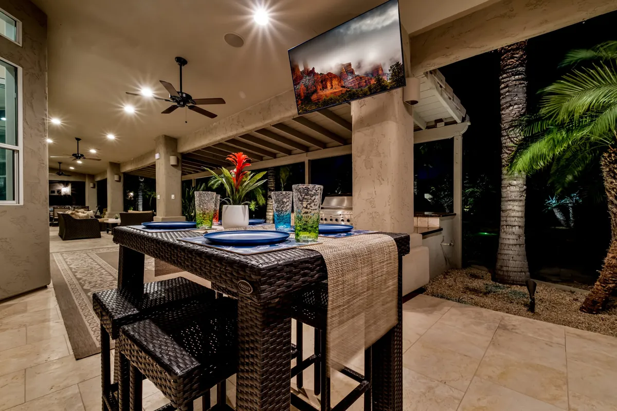 A festive exterior dining scene with a dining table set with colorful plates and glasses, tropical plants, a TV displaying a scenic image, and palm trees in the background. The setup suggests a space for group dining, outdoor movie nights, and casual celebrations under the stars.