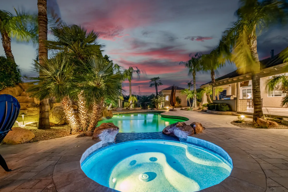 This image displays a serene backyard setting featuring a heated swimming pool, adjacent hot tub, lush palm trees, and meticulously landscaped surroundings. The sky glows with sunset colors, adding a touch of luxury and tranquility. The pool and spa are focal points, inviting relaxation and play, while the overall scene feels private and secluded.