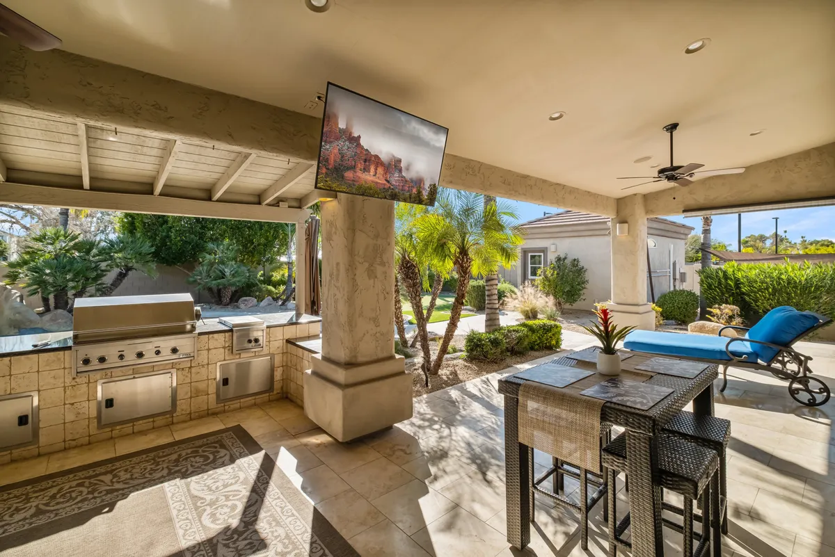 This image highlights a stylish outdoor kitchen with a built-in grill, a dining area, a comfortable lounge chair, a flat-screen patio TV, and lush landscaped gardens. The setup is ideal for grilling, al fresco dining, and watching movies outdoors, with the garden and lighting creating an inviting atmosphere.