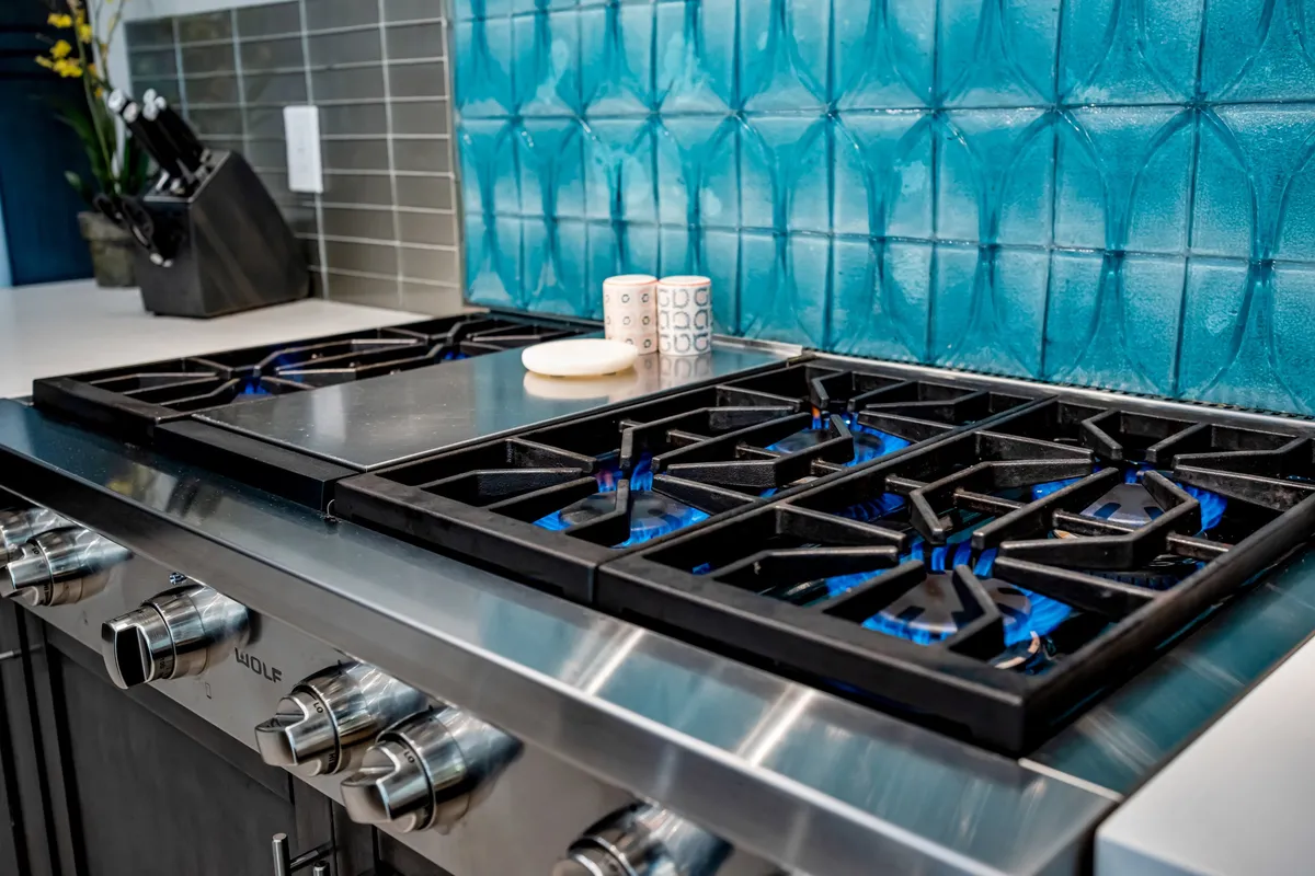 A close-up view of the chef’s kitchen highlights a gleaming stainless steel gas stove, complemented by a textured teal backsplash and neatly arranged utensils. The scene is lively and energetic, evoking the spirit of group cooking and culinary creativity.